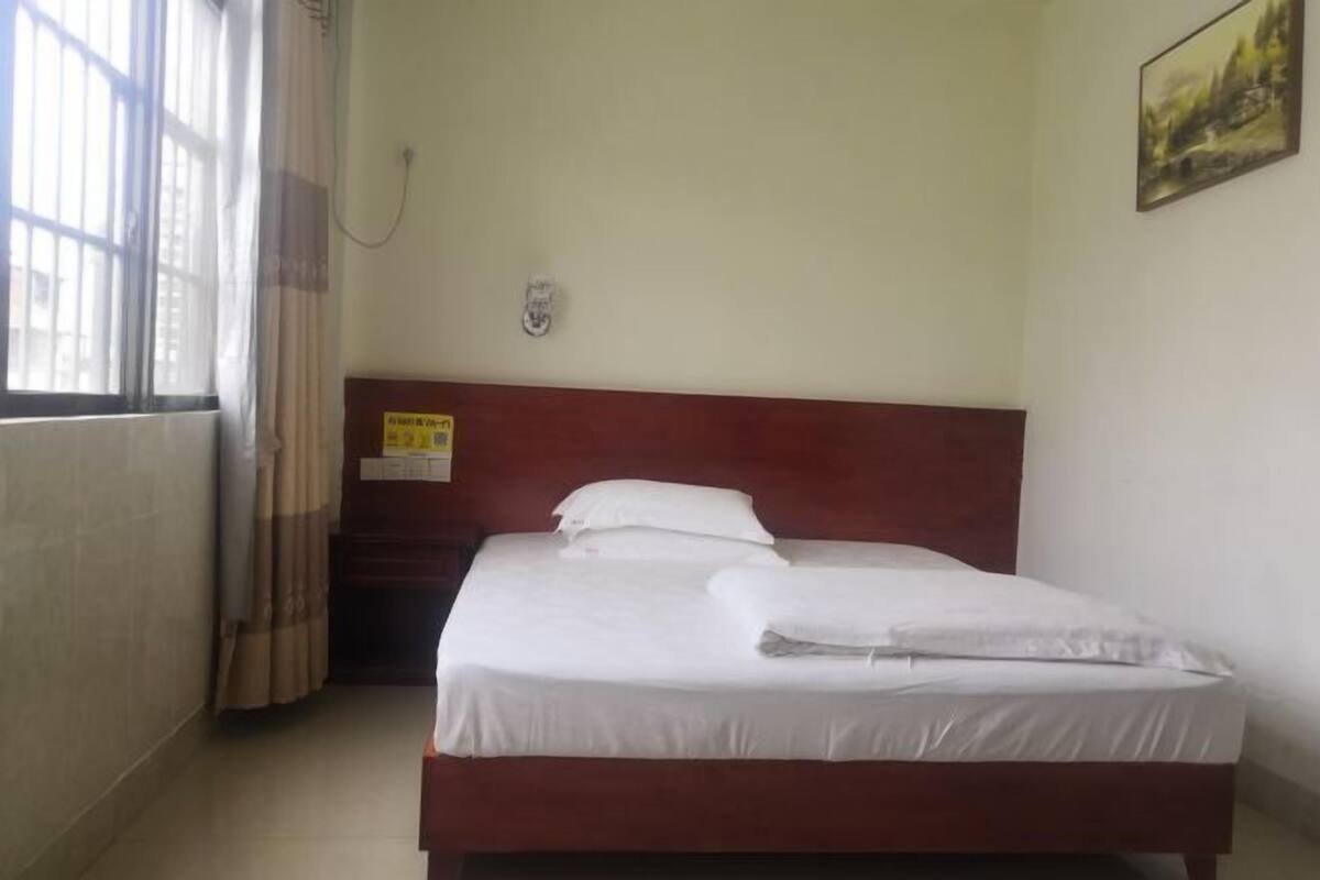 Una cama individual con cabecera de madera, una ventana con cortinas, y un aplique de pared.