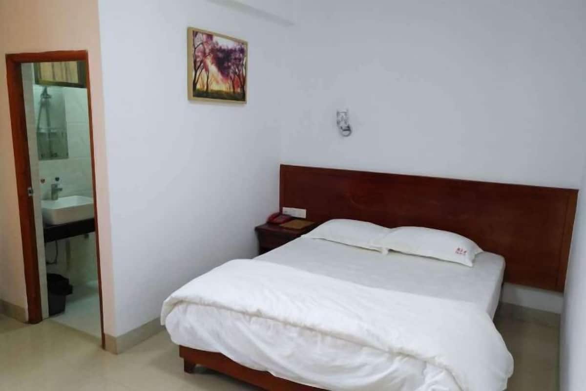Un dormitorio con cama, una mesita de noche, una lámpara de pared, un cuadro y un baño visible a través de una puerta abierta.