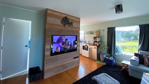 Ein modernes Wohnzimmer mit einem Flachbildfernseher an einer Holzwand, einer Küchenzeile mit Mikrowelle und Herd sowie einem großen Fenster mit Blick auf Grünflächen.