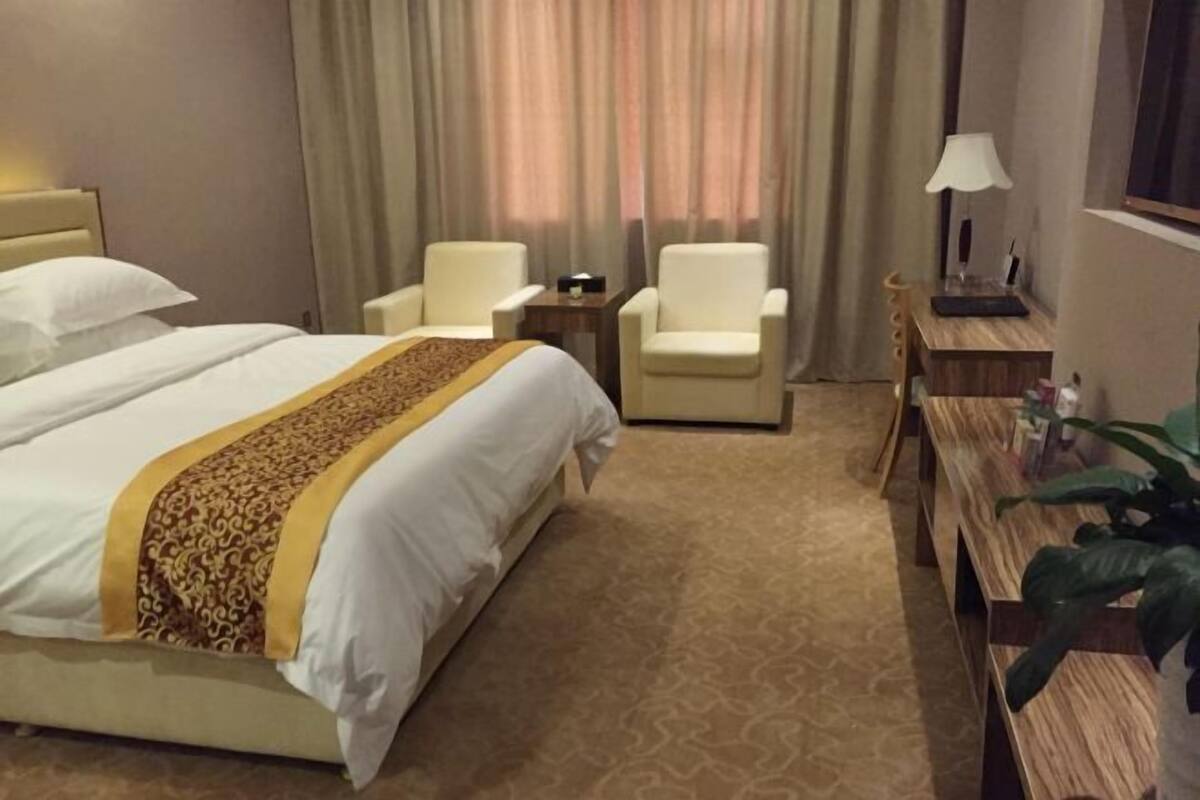 Een hotelkamer met een groot bed, twee fauteuils, een klein tafeltje, een lamp en een houten bankje.