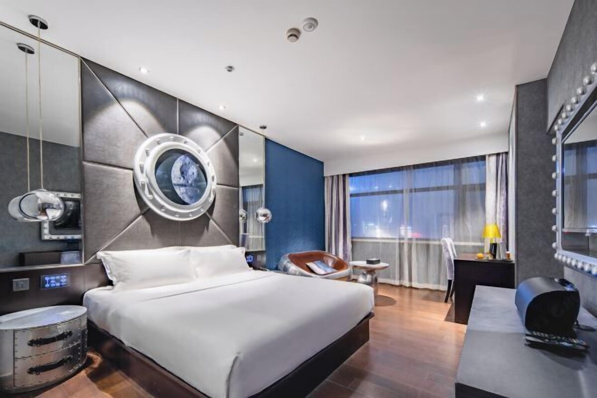 Quarto de hotel moderno com uma cama grande, uma toucador com espelho, um espaço de estar e uma janela ampla.