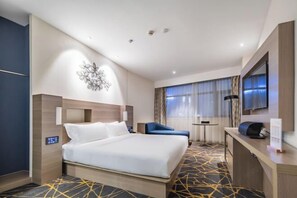 Quarto de hotel com cama, escrivaninha, cadeira, uma obra de arte na parede e janela com cortinas.