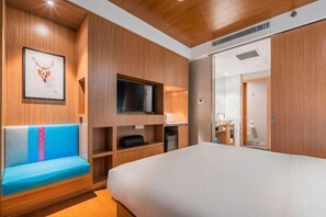 Quarto de hotel moderno com uma cama grande, TV embutida, área de estar com um almofão azul e painéis de madeira.