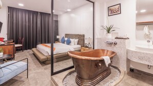 A modern hotel room with a large bed, a sitting area with a chair and table, a unique copper bathtub, and a bathroom with a large mirror and sink. - Bamboo Retreat Villa (Siem Reap)