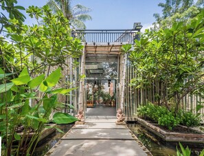 A pathway leading to a building with a glass entrance, flanked by lush greenery and two stone statues. - Bamboo Retreat Villa (Siem Reap)