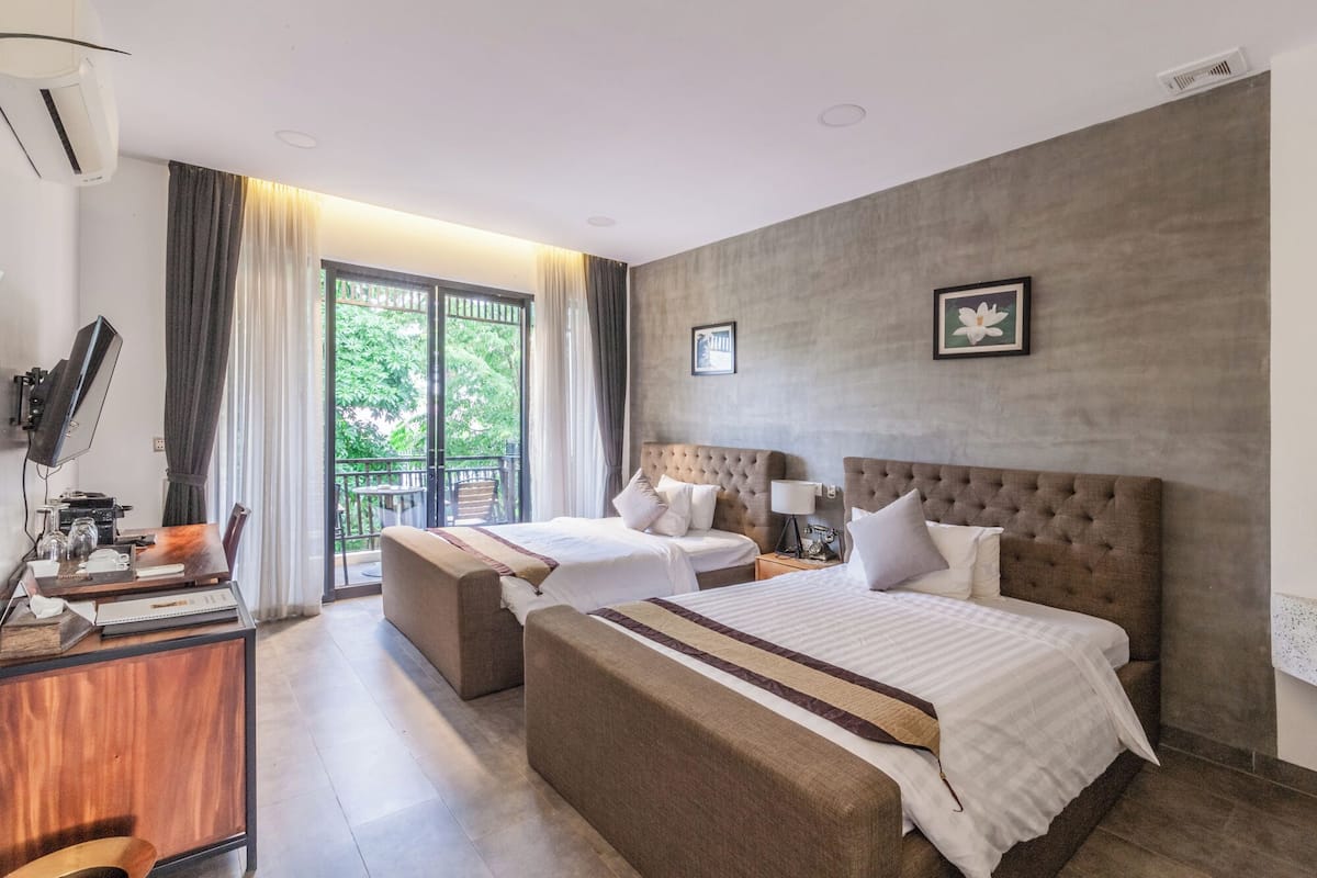 A hotel room with two beds, a desk with a computer, and a balcony with a view of greenery.