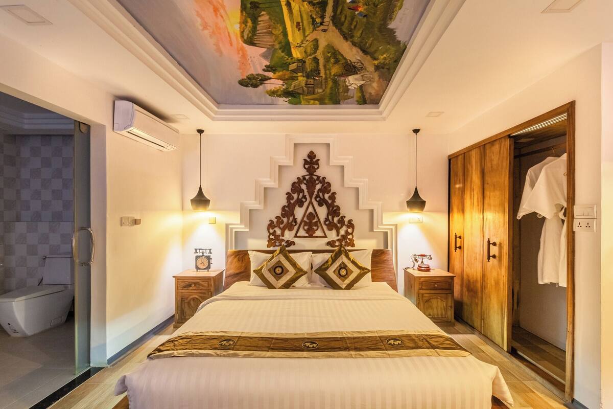 A hotel room with a bed, a wooden wardrobe, a ceiling mural, and a nightstand with a clock.