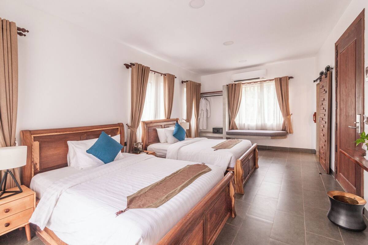 A hotel room with two wooden beds, beige curtains, a wooden wardrobe, and a window with a view.