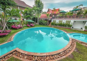 A swimming pool with lounge chairs and umbrellas, surrounded by greenery and a building with a red roof. - Bamboo Retreat Villa (Siem Reap)