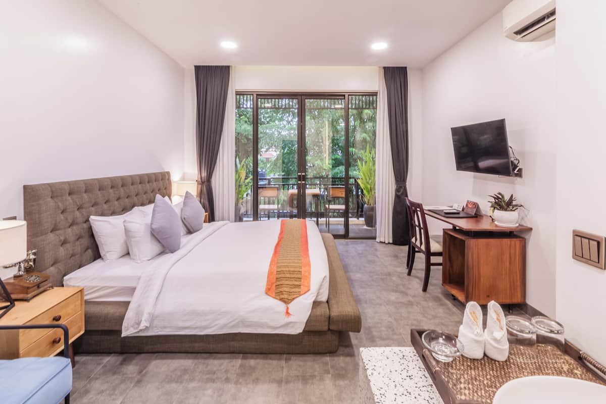 A hotel room with a bed, a desk, a chair, a TV, and a balcony with a view of greenery.