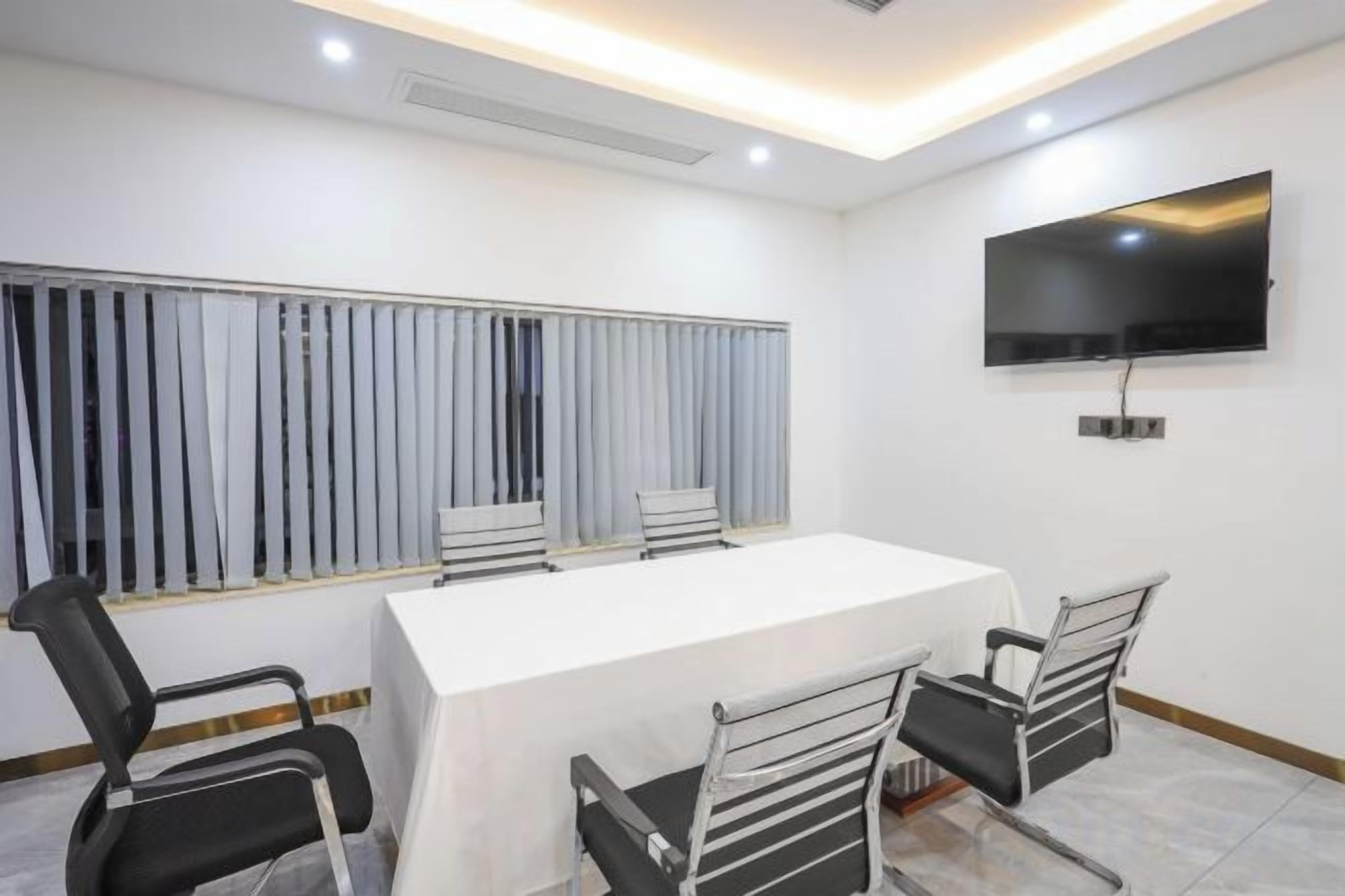A conference room with a long table, four chairs, a TV on the wall, and blinds on the window.