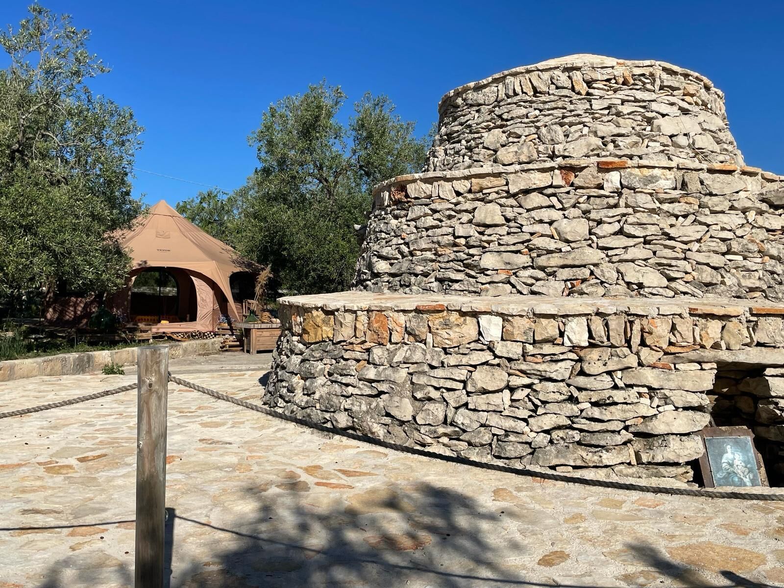 Una estructura de piedra con parte superior circular, posiblemente un sitio histórico o cultural, rodeada por un área pavimentada y un edificio tipo tienda de campaña al fondo.