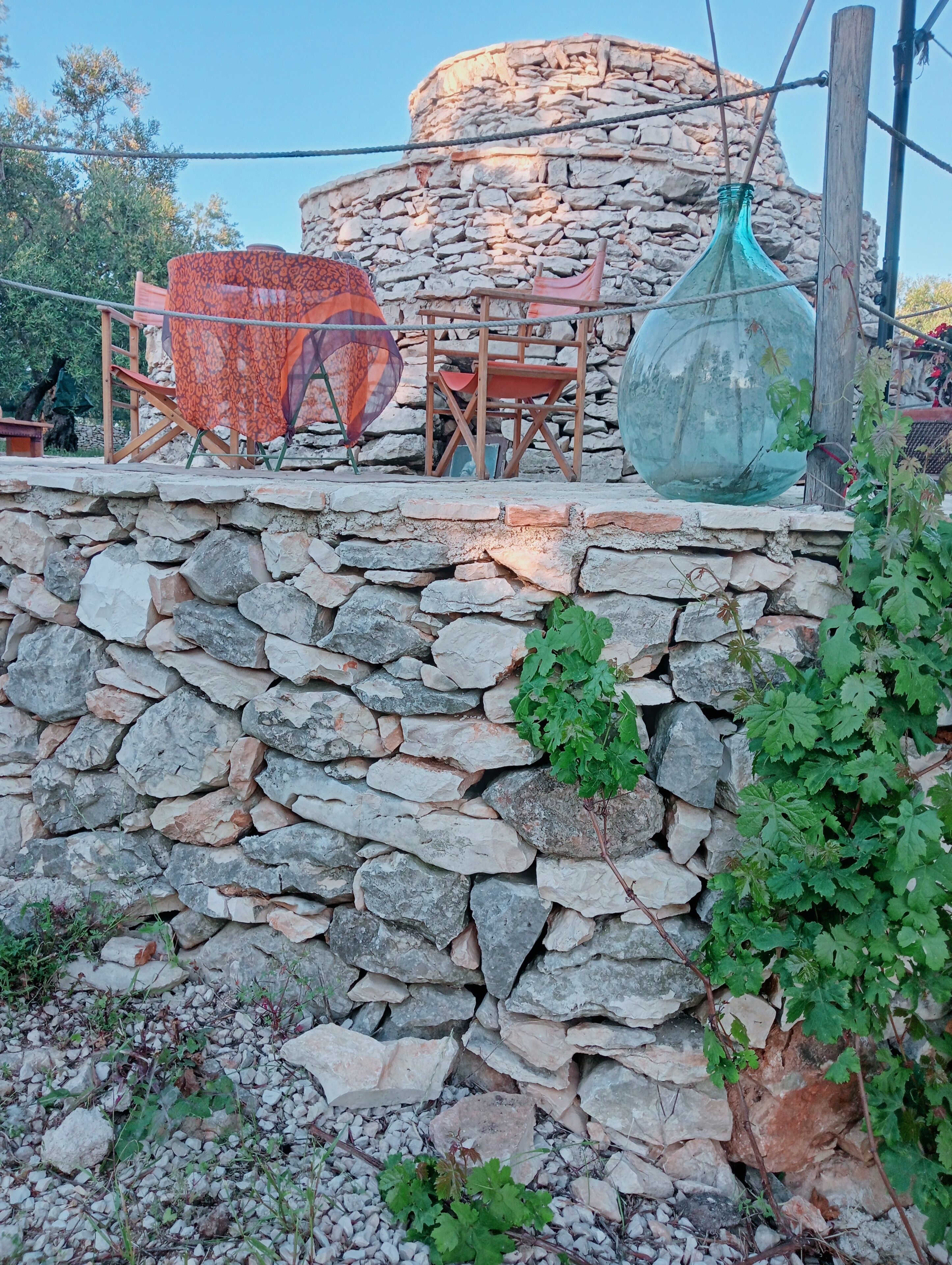 Paret de pedra amb una estructura de fusta rústica a la part superior, que inclou una cadira de fusta i una ampolla de vidre gran.
