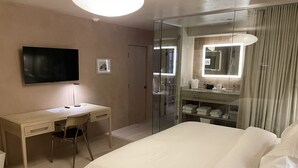 A hotel room with a bed, a desk, a chair, a TV, a glass shower enclosure, and a bathroom with a mirror and sink.