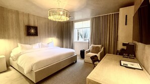 A hotel room with a large bed, a desk, a chair, a small table, a lamp, and a window with curtains. - Palace Modern Hotel at the Plaza - Premium King 8 (Santa Fe)
