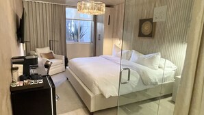 Una habitación de hotel moderna con una cama grande, un separador de vidrio, una mesita de noche con artículos de cortesía, un sofá y un televisor.