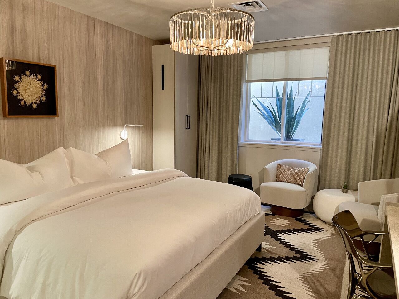 A modern hotel room with a large bed, a sitting area with a chair and ottoman, a chandelier, and a window with curtains.