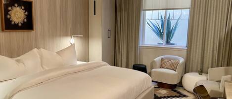 A modern hotel room with a large bed, a sitting area with a chair and ottoman, a chandelier, and a window with curtains.