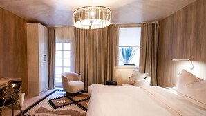 Una camera d'albergo con un letto grande, una scrivania con una sedia, una poltrona e un lampadario, oltre a una finestra con tende.