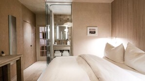 Une chambre moderne avec un grand lit, une table de chevet, un miroir et une salle de bain visible à travers une porte en verre.