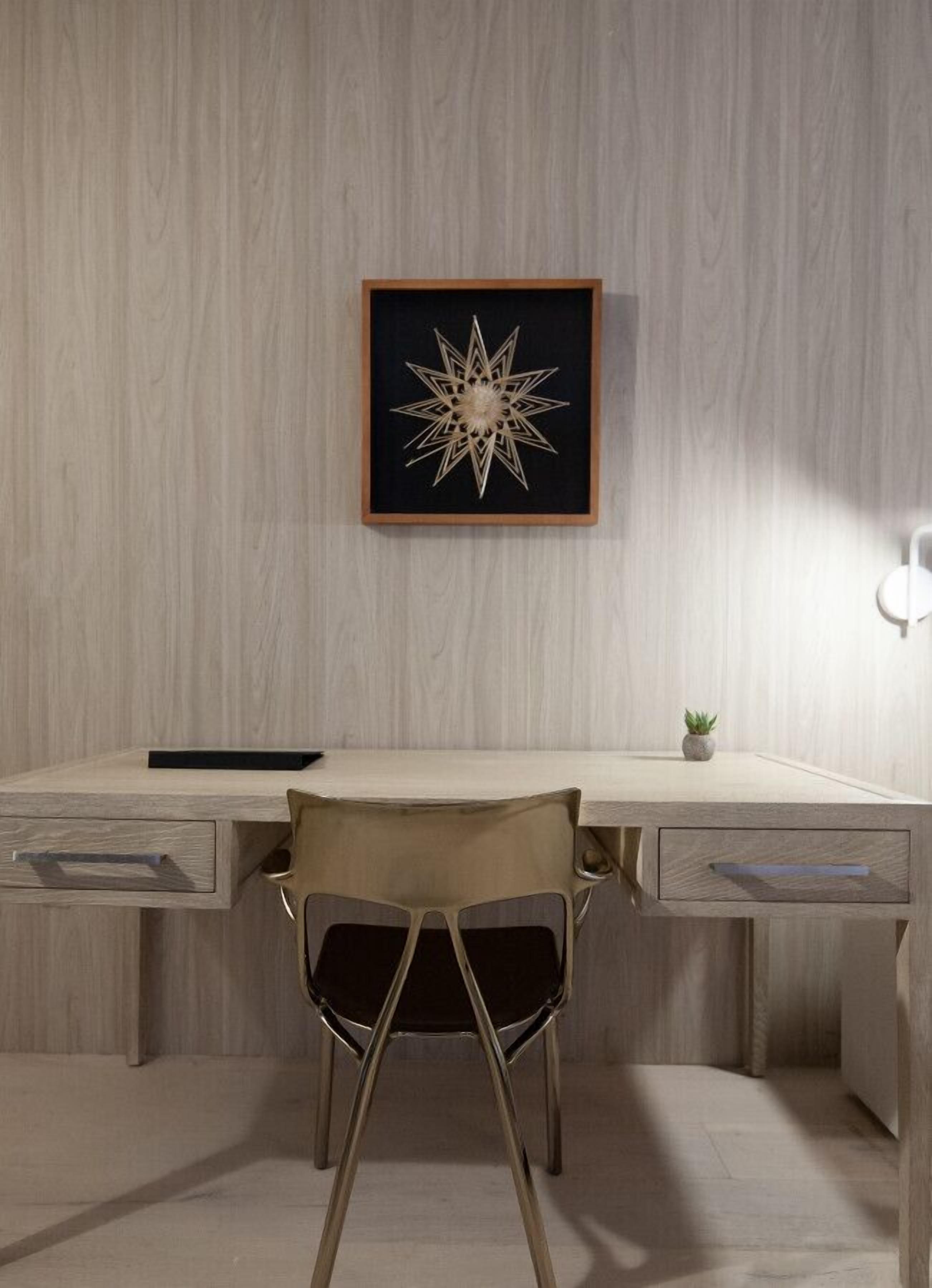 Un escritorio moderno con una obra de arte geométrica enmarcada, una silla y una planta pequeña sobre el escritorio.