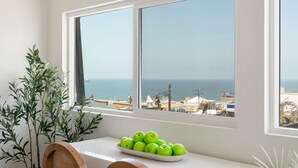 Ein Zimmer mit großem Fenster mit Blick aufs Meer, ein Tisch mit einer Schüssel grüner Äpfel und eine Pflanze neben dem Fenster.