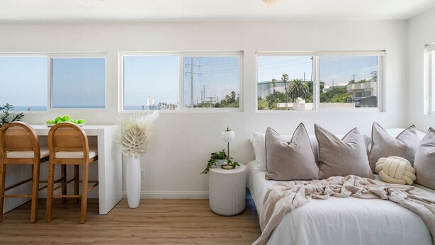 Ein modernes Schlafzimmer mit einem Bett, einem Schreibtisch und zwei Stühlen. Große Fenster bieten Blick auf das Meer und Gebäude.