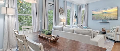 A dining area with a wooden table, white chairs, and a view of greenery outside.