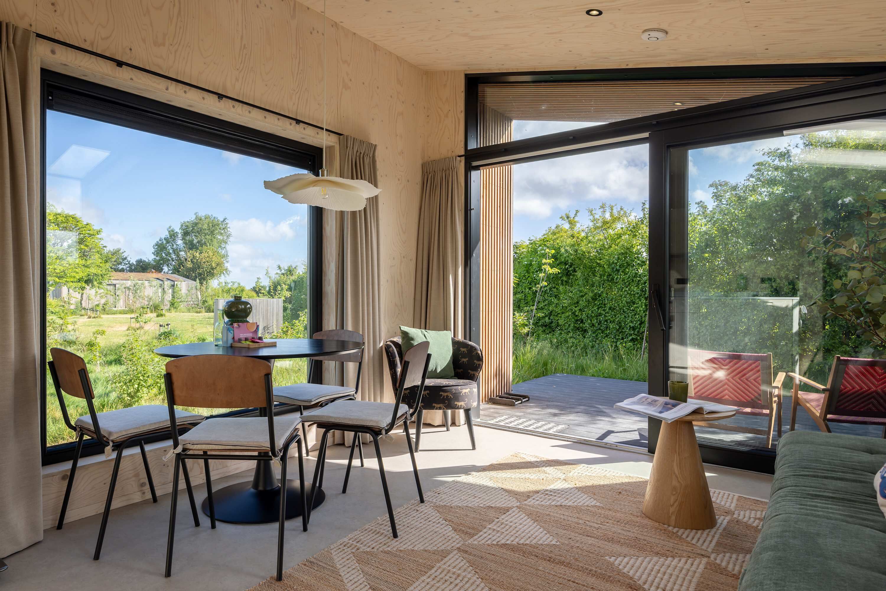 A modern interior with a dining area, a sofa, and large windows offering a view of greenery and a distant building.