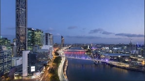 Eine Stadtansicht im Dämmerlicht mit modernen Gebäuden, einem Fluss und einer Brücke mit bunten Lichtern.