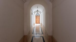 Un pasillo con una puerta de madera al final, paredes blancas y suelo de mármol.