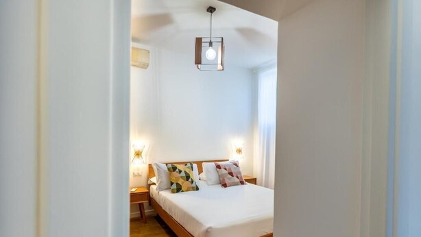 Un dormitorio con cama de madera, ropa de cama blanca y almohadas con estampado geométrico. Hay lámparas de noche y una lámpara colgante.