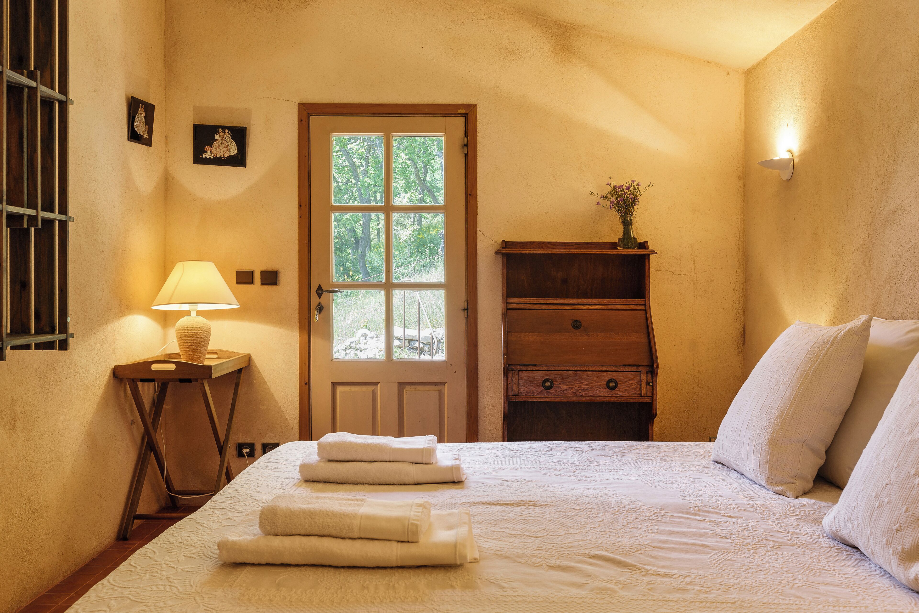 A bedroom with a bed, a nightstand with a lamp, a wooden dresser, and a door with a window.
