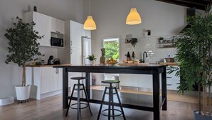 A modern kitchen with white cabinets, a central island with stools, pendant lighting, and a view of greenery outside. - Amazing 180° Ocean View 3 Bedroom Villa w/ Swimming Pool  (Capelo - Horta)