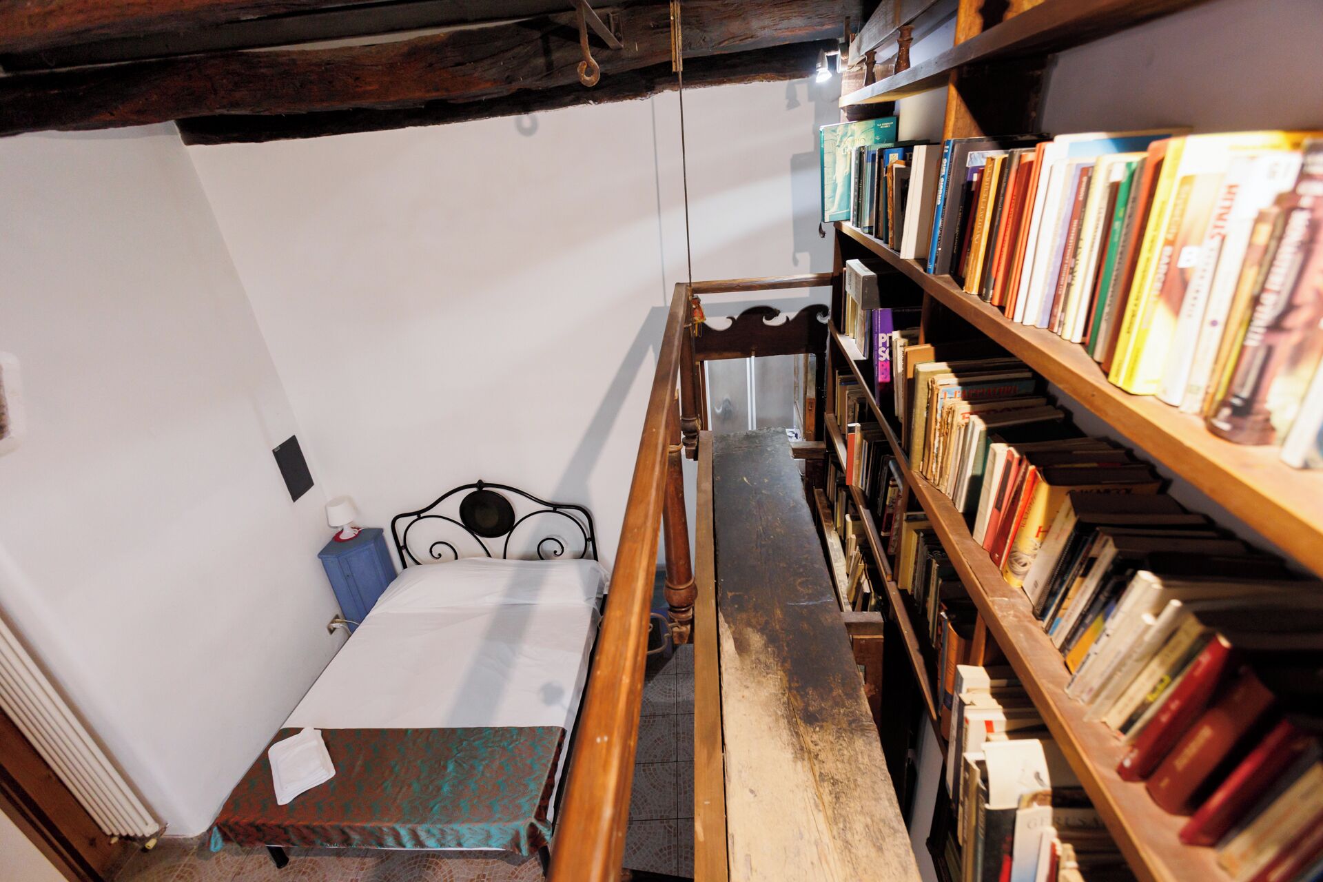 A narrow staircase leading to a bedroom with a wooden headboard and a bedside table. A bookshelf filled with books is visible to the right.
