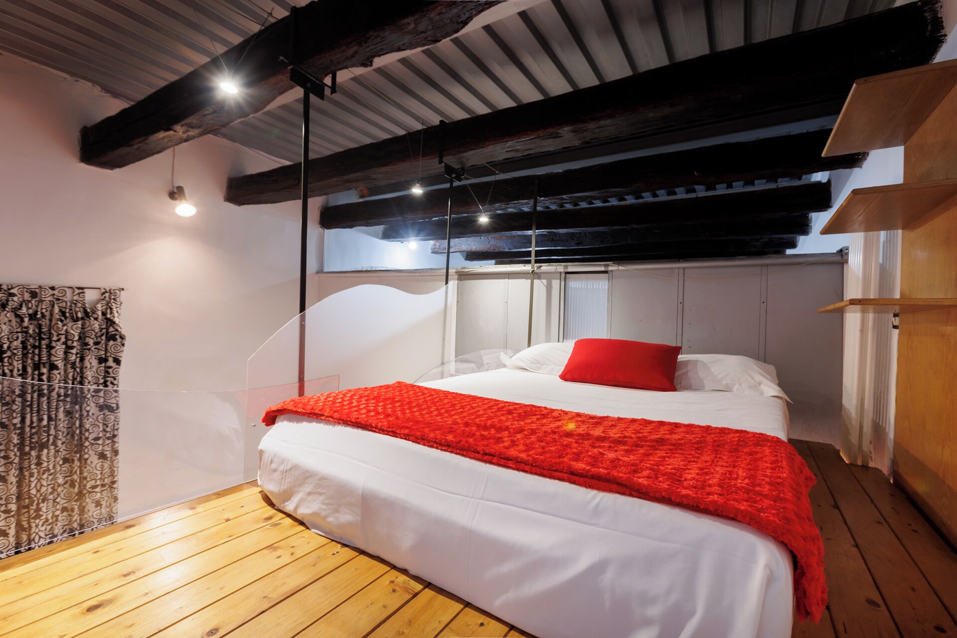 A bedroom with a wooden floor, a bed with white bedding and a red throw, a wooden ceiling, and a window with curtains.