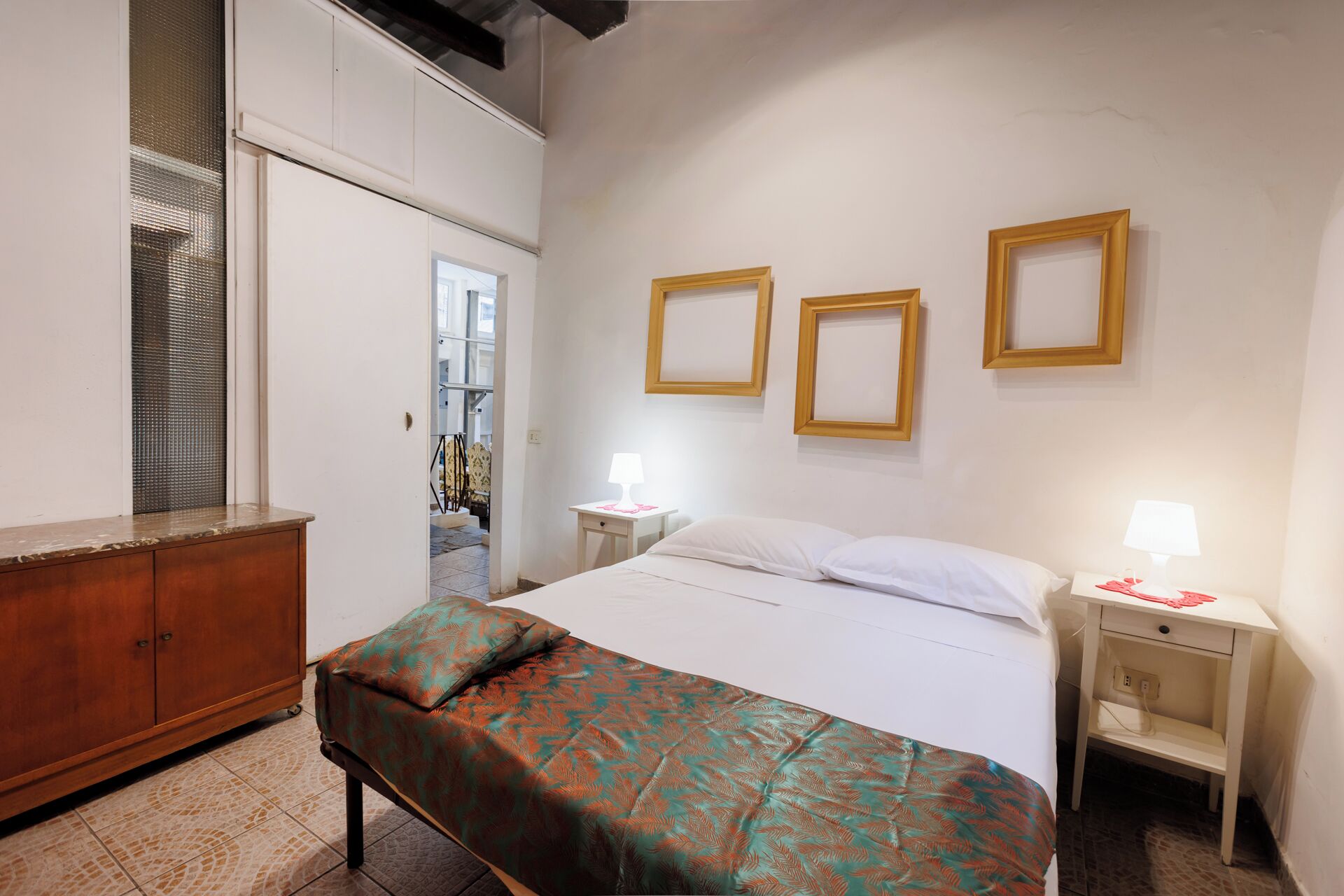 A hotel room with a bed, bedside tables, a bench, a wardrobe, and framed pictures on the wall.