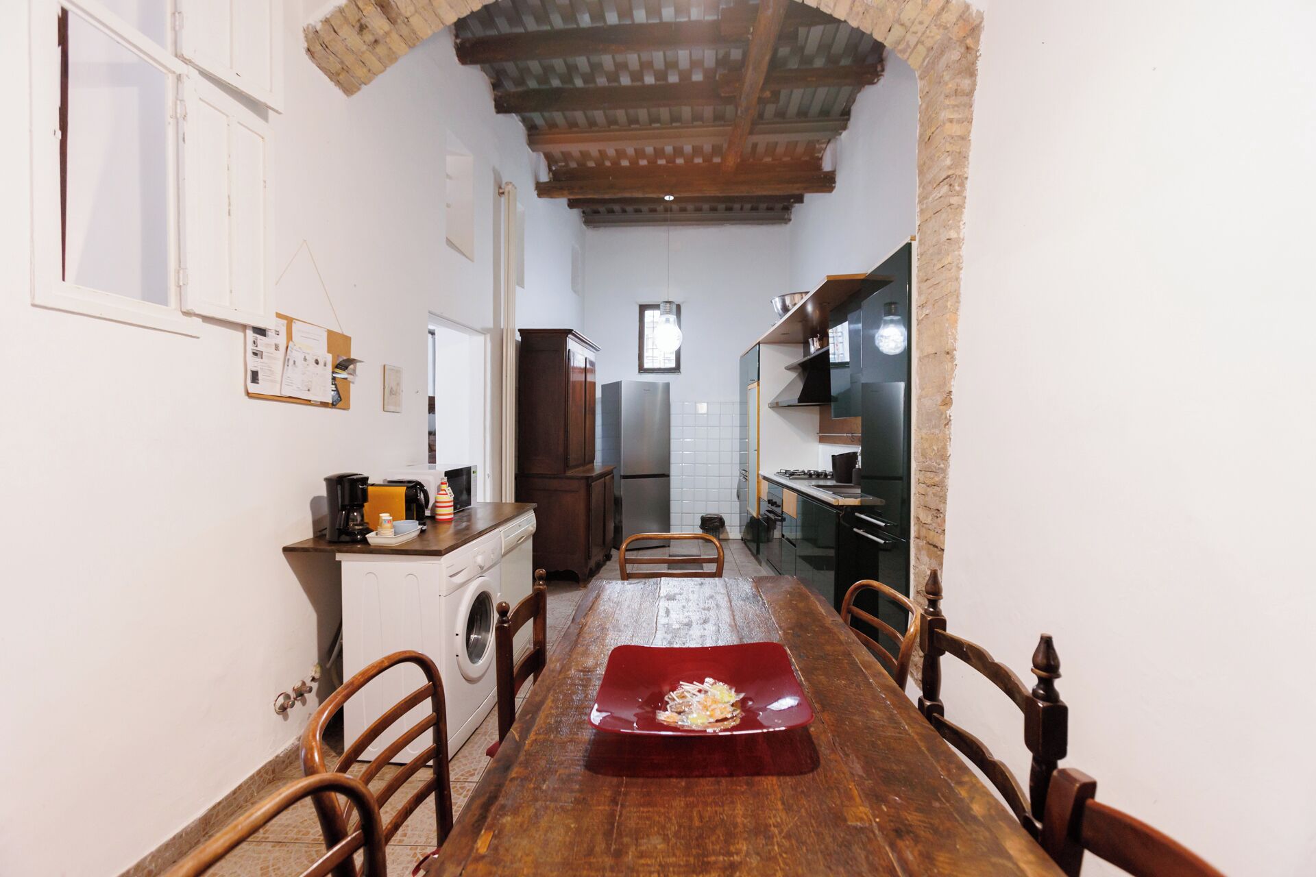 A narrow kitchen with a wooden table and chairs, a washing machine, and a refrigerator.