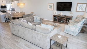 A modern living room with a large sofa, a coffee table, and a TV. There is a kitchen in the background with a dining area. - 3 br, gulf view, beach club, pools, gym, hot tub (Pensacola)