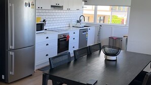 A modern kitchen with white cabinets, a stainless steel refrigerator, a microwave, and a dishwasher. There is a dining table with black chairs in the foreground. - Beach View Cottage. Cosy with direct water views. (Clontarf)