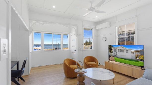 A spacious living room with a large window, a ceiling fan, and a television displaying a house. - Beach View Cottage. Cosy with direct water views. (Clontarf)