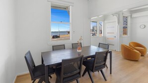 A modern dining area with a large table, six chairs, and a vase with flowers. A window offers a view of the street and buildings. - Beach View Cottage. Cosy with direct water views. (Clontarf)