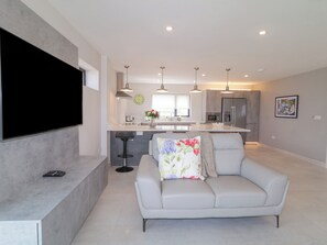 A modern living room with a flat-screen TV, a grey sofa with floral cushions, and a kitchen in the background with pendant lighting.