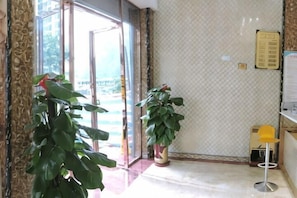 A hotel lobby with a glass door entrance, potted plants, a yellow chair, and a bulletin board. - Taijinyun Business Hotel (Hechi)