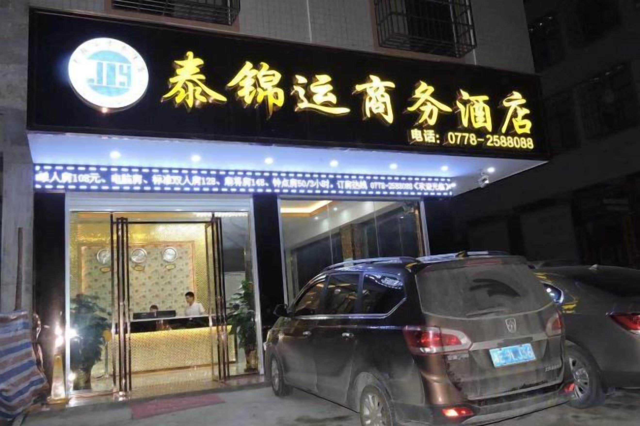 A hotel entrance with a reception desk, a car parked in front, and a signboard with contact information.