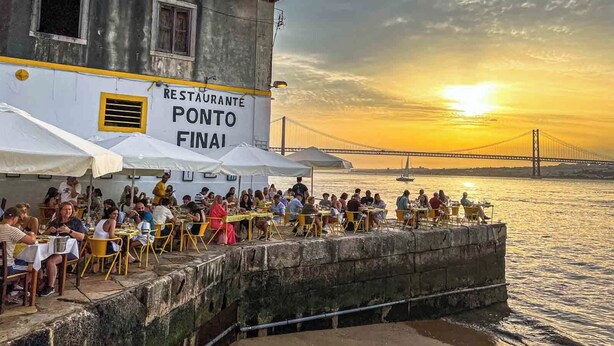 A waterfront restaurant named 'RESTAURANTE PONTO FINAI' with outdoor seating and a view of a bridge in the background. - Over the Rainbow  (Almada)