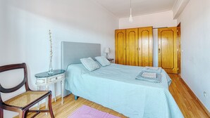 A bedroom with a bed, wooden wardrobe, a bedside table with a lamp, and a chair. - Over the Rainbow  (Almada)