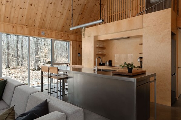 Een moderne keuken met een centraal eiland, houten plafond en grote ramen met uitzicht op een bos.