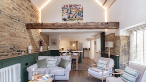 A cozy living room with a stone wall, wooden beams, a dining area, and a kitchen in the background. - A spectacular barn conversion - Highgate (Moreton-in-Marsh)