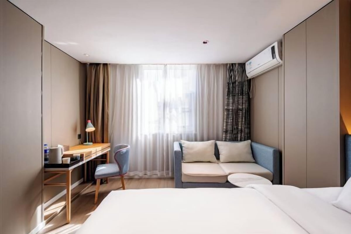 A hotel room with a bed, a desk with a chair, a sofa, and a window with curtains.